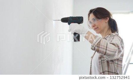 Middle aged happy woman wearing protective gear and gloves, is drilling hole in white wall during home improvement project, focused on precise home renovation work Middle aged happy woman wearing protective gear and gloves, is drilling hole in white wall during home improvement project, focused on precise home renovation work 132752941