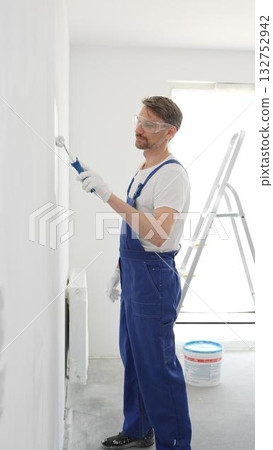 Middle aged construction worker in a blue jumpsuit, safety goggles, and work gloves, applying paint with roller on pristine interior wall during home renovation project, vertical view Middle aged construction worker in a blue jumpsuit, safety goggles, and work gloves, applying paint with roller on pristine interior wall during home renovation project, vertical view 132752942