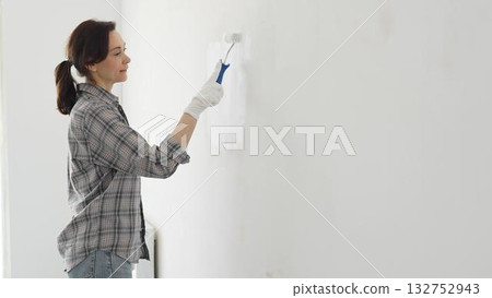 Middle aged woman wearing plaid shirt and white gloves is painting white wall with paint roller, experiencing home renovation and transforming living space. Home repair concept Middle aged woman wearing plaid shirt and white gloves is painting white wall with paint roller, experiencing home renovation and transforming living space. Home repair concept 132752943