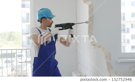Male construction worker demolishing concrete wall with rotary hammer, wearing work overalls and blue hardhat is generating dust and debris during home renovation work, portrait horizontal view Male construction worker demolishing concrete wall with rotary hammer, wearing work overalls and blue hardhat is generating dust and debris during home renovation work, portrait horizontal view 132752946