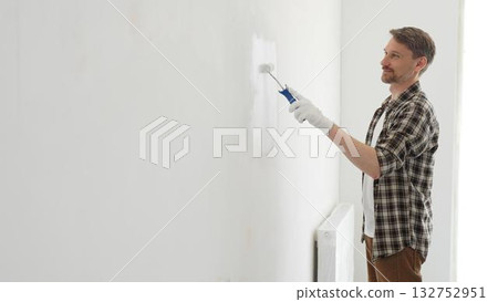 Middle aged man wearing checked shirt and protective gloves, is painting a white wall with a paint roller, working on a home renovation project, creating a fresh and clean look Middle aged man wearing checked shirt and protective gloves, is painting a white wall with a paint roller, working on a home renovation project, creating a fresh and clean look 132752951