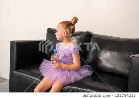 Girl ballerina with cochlear implant sitting on black sofa holding bottle of water. Inclusion, childhood recovery, and empowerment through hearing and rest. 132754216