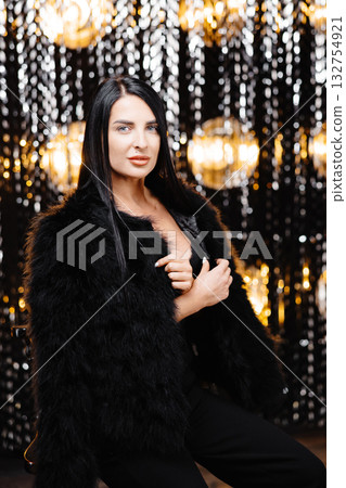 Young beautiful smiling brunette female in trendy evening fur coat. Carefree woman posing near shiny tinsel wall in studio. Fashionable model with bright makeup looks at camera Young beautiful smiling brunette female in trendy evening fur coat. Carefree woman posing near shiny tinsel wall in studio. Fashionable model with bright makeup looks at camera 132754921