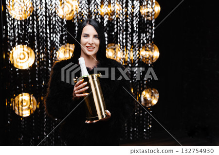Charming woman with brunette long hair in trendy evening fur coat holding bucket with bottle with champagne on shine wall in studio 132754930
