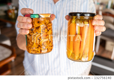 Caucasian adult holding jars of pickled carrots and mushrooms in kitchen setting. National Pickle Day Caucasian adult holding jars of pickled carrots and mushrooms in kitchen setting. National Pickle Day 132756140