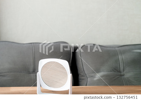 A white round plastic makeup mirror stands on a wooden table against the background of a sofa 132756451