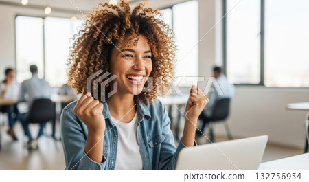 Young woman with curly hair is celebrating success while working on a laptop in a modern office environment, showcasing joy and achievement 132756954