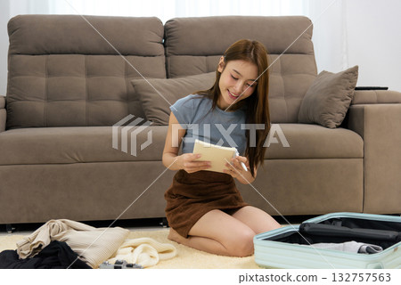 Happy young woman home activity, people planning trip, writing essential items in notebook, packing open suitcase with clothes for vacation preparation. Happy young woman home activity, people planning trip, writing essential items in notebook, packing open suitcase with clothes for vacation preparation. 132757563