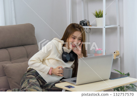 Smiling young happy woman enjoys home activity, relaxing on comfortable sofa browsing laptop, holding warm mug, wearing headphones for casual digital entertainment. Smiling young happy woman enjoys home activity, relaxing on comfortable sofa browsing laptop, holding warm mug, wearing headphones for casual digital entertainment. 132757570