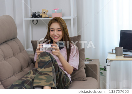Young happy woman at home enjoying gaming activity, people embracing leisure lifestyle, relaxing indoor pastime, holding white controller, wearing camouflage pants, headphones around neck. 132757603