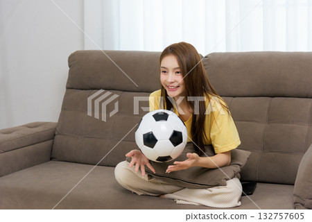 Young happy woman at home enjoying sport activity, sitting comfortably on cozy sofa, holding soccer ball, eagerly anticipating game with bright smile, showing cheerful spirit. 132757605