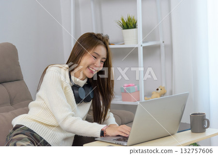 Young happy woman enjoys home activity, smiling broadly while using laptop computer for remote work or online learning, headphones nearby, modern lifestyle. 132757606