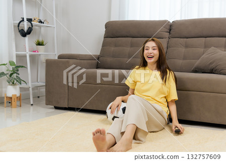 Young woman home happy activity watching television program, enjoying leisure time indoors, smiling with remote control, soccer ball near, relaxing in comfortable living room. Young woman home happy activity watching television program, enjoying leisure time indoors, smiling with remote control, soccer ball near, relaxing in comfortable living room. 132757609