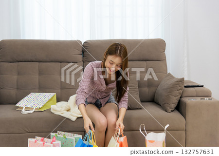 Young woman home activity, happy people enjoying new purchases, colorful shopping bags spread around sofa, smiling lady carefully unpacking delightful finds after successful shopping trip at home. 132757631