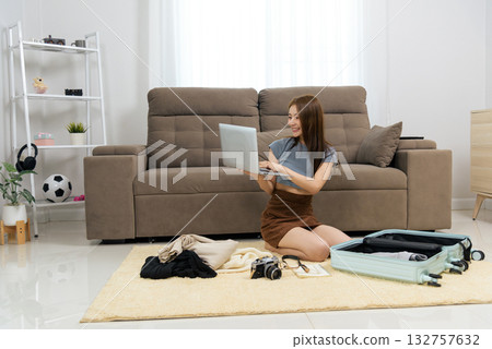 Young woman happy home activity people enjoy, sitting on rug, using laptop for travel planning, packing suitcase, arranging clothing, camera and notebook ready for vacation. Young woman happy home activity people enjoy, sitting on rug, using laptop for travel planning, packing suitcase, arranging clothing, camera and notebook ready for vacation. 132757632