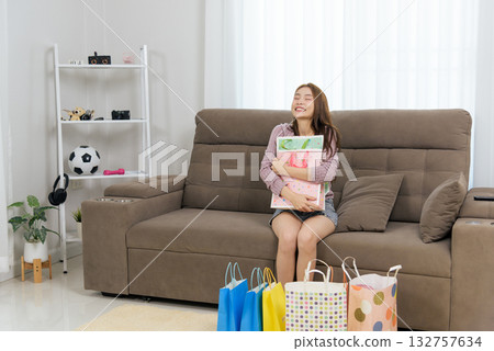 Young happy woman home shows people joy, enjoying leisure activity, embracing colorful gift bags, eyes closed in delight, sitting comfortably on sofa, surrounded by bright shopping bags on floor. Young happy woman home shows people joy, enjoying leisure activity, embracing colorful gift bags, eyes closed in delight, sitting comfortably on sofa, surrounded by bright shopping bags on floor. 132757634
