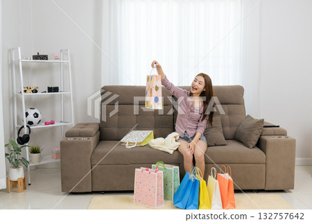 Young happy woman at home engages in activity people enjoy, admiring shopping purchases on sofa surrounded by many colorful bags. Young happy woman at home engages in activity people enjoy, admiring shopping purchases on sofa surrounded by many colorful bags. 132757642