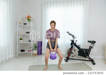 Young smiling woman performs fitness activity at home, exercising with purple kettlebell, training for healthy body barefoot on yoga mat in home gym room. 132757643