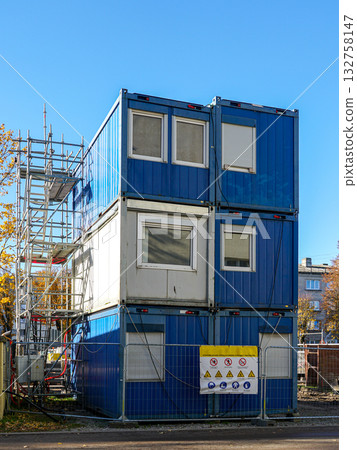 Stacked blue modular office containers at construction site with safety sign and scaffolding 132758147