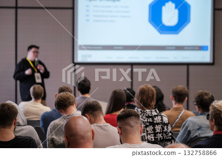 Presenter delivers talk to engaged crowd in conference room with large projection screen Presenter delivers talk to engaged crowd in conference room with large projection screen 132758866