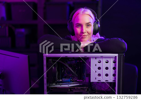 Female gamer with headphones leans on an open gaming PC case under purple lighting, ready to upgrade hardware 132759286