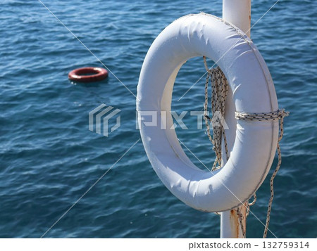White Lifebuoy On Pole Over Blue Water With Red Buoy In Background - Nautical Safety Image White Lifebuoy On Pole Over Blue Water With Red Buoy In Background - Nautical Safety Image 132759314