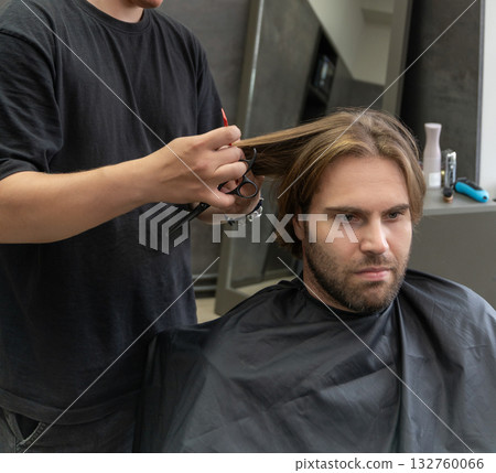 A hairdresser uses a pair of scissors to cut a male client's long blonde hair. 132760066