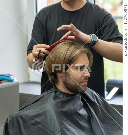 A hairdresser uses a pair of scissors to cut a male client's long blonde hair. A hairdresser uses a pair of scissors to cut a male client's long blonde hair. 132760071