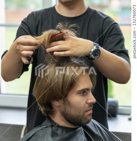 A hairdresser uses a pair of scissors to cut a male client's long blonde hair. A hairdresser uses a pair of scissors to cut a male client's long blonde hair. 132760073