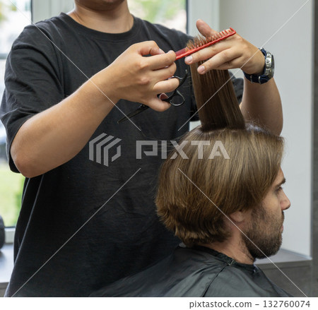 A hairdresser uses a red comb and scissors to style a male client's long brown hair. 132760074