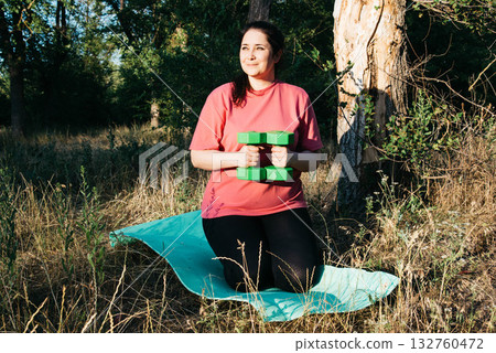 A plus-size woman doing dumbbell exercises outdoors. Outdoor sports. Green dumbbells in the athlete's hands. High quality photo 132760472