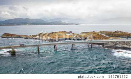Dramatic engineering of Atlantic ocean road weaves across small rugged islands. Road stretches among calm waters to foggy distance under overcast sky Dramatic engineering of Atlantic ocean road weaves across small rugged islands. Road stretches among calm waters to foggy distance under overcast sky 132761252