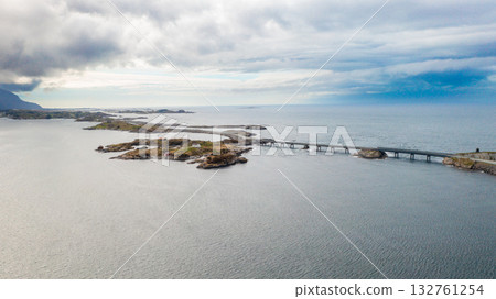 Magnificent stretch of Atlantic ocean road with long bridge gracefully spanning deep blue waters. Rough waves crash near rugged coastline in Norway 132761254
