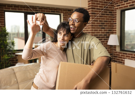 Diverse couple presenting house keys and carrying cardboard box during relocation. Boyfriend and girlfriend celebrate becoming homeowners and new beginnings in modern apartment on moving day. Diverse couple presenting house keys and carrying cardboard box during relocation. Boyfriend and girlfriend celebrate becoming homeowners and new beginnings in modern apartment on moving day. 132761632