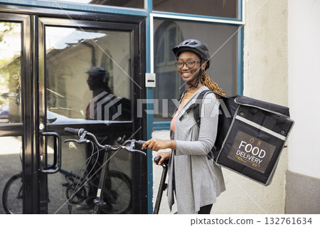 Portrait of african american woman working as a courier with bike, carrying thermal backpack to bring fast food orders to clients. Riding the delivery bike with helmet and takeaway bag. Portrait of african american woman working as a courier with bike, carrying thermal backpack to bring fast food orders to clients. Riding the delivery bike with helmet and takeaway bag. 132761634
