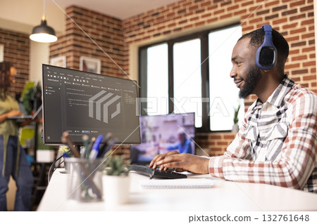 Black male programmer smiling at desktop monitor and reviewing software code. Confident developer working remotely on secure website for startup tech company from home office. 132761648