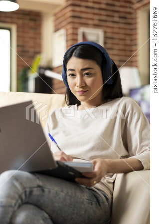 Remote female manager preparing for virtual meeting, holding pen while reviewing marketing notes. Asian woman entrepreneur with headphones and notebook, seated on sofa, focusing on strategic planning. 132761669