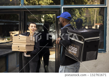 Black courier handing a heavy pile of pizza boxes to a young woman, express takeout service. Male employee brings takeaway meal from a fast food restaurant in an urban setting. 132761671
