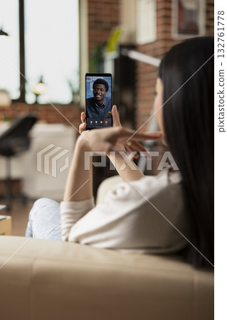 Remote female manager seated on sofa, holding smartphone during video call, chatting with black male colleague. Businesswoman engaged in mobile online meeting with african american investor. Remote female manager seated on sofa, holding smartphone during video call, chatting with black male colleague. Businesswoman engaged in mobile online meeting with african american investor. 132761778