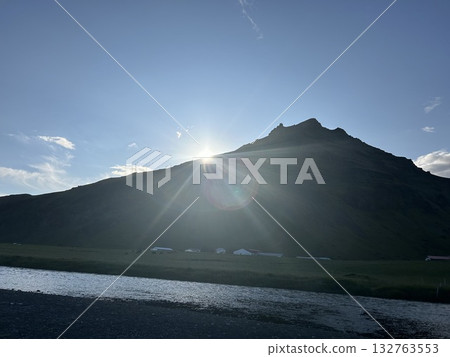 A river beyond the waterfall, the sun, and the green moss-covered mountains that line Skógafoss waterfall in southern Iceland. 132763553