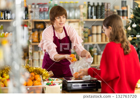 Positive adult woman seller in grocery store standing behind counter full of products for Christmas table and weighing and selling tangerines to girl Positive adult woman seller in grocery store standing behind counter full of products for Christmas table and weighing and selling tangerines to girl 132763720