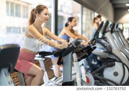 Smiling young girl doing spin bike exercises in well-equipped gym Smiling young girl doing spin bike exercises in well-equipped gym 132763792