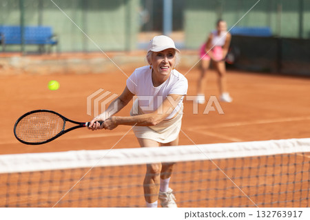 Sporty two women during doubles match on tennis court. Active old lady hitting tennis ball with racket Sporty two women during doubles match on tennis court. Active old lady hitting tennis ball with racket 132763917