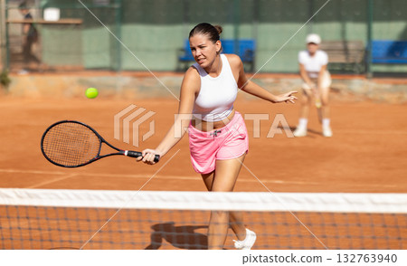 Active young girl hits tennis ball with racket in front of players in court. Two sportive women enjoying padel tennis Active young girl hits tennis ball with racket in front of players in court. Two sportive women enjoying padel tennis 132763940
