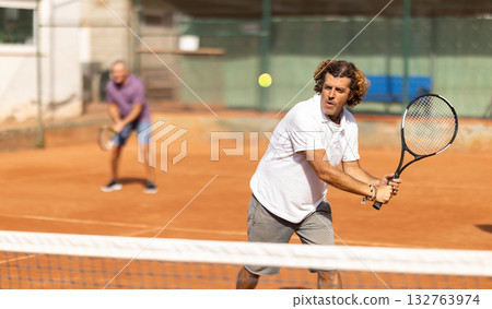 Sporty two men during doubles match on tennis court. Active sportsman hitting tennis ball with racket Sporty two men during doubles match on tennis court. Active sportsman hitting tennis ball with racket 132763974