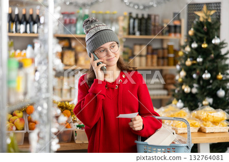 She holds paper note in hands, asks mother questions on phone while shopping in grocery 132764081