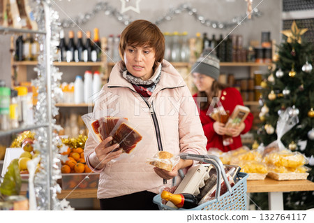 Positive woman with shopping cart or basket choosing dried meat or jamon for New Year's holiday in supermarket Positive woman with shopping cart or basket choosing dried meat or jamon for New Year's holiday in supermarket 132764117