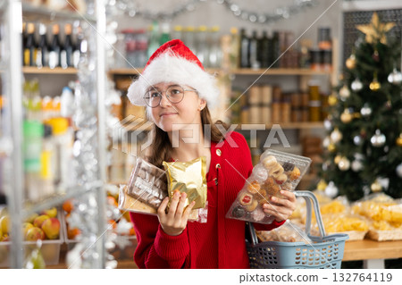 Portrait of teenage girl with candies and different sweets in her hands in supermarket decorated with tinsel and Christmas tree 132764119