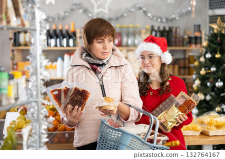 Adult woman and girl in festive hat looking for something, holding cart with food products at store. Preparation for New Year celebration 132764167