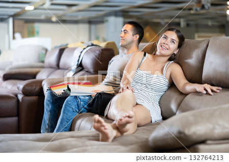 In store wife settled down with legs to test of sofa in use,husband examines upholstery fabric 132764213